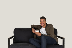 Front view of a smiling young man sitting on a sofa with a cup of coffee