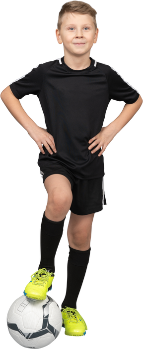Vista frontale di un ragazzo bambino sorridente in uniforme di calcio mettendo le mani sui fianchi e il piede sulla palla