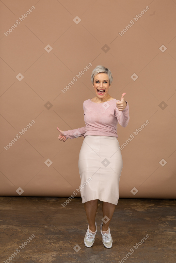 Vista frontal de una mujer feliz en ropa casual de pie sobre los dedos de los pies y mostrando el pulgar hacia arriba