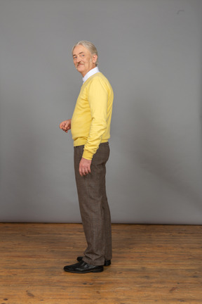 Side view of a smiling old man in yellow pullover raising hand and looking aside