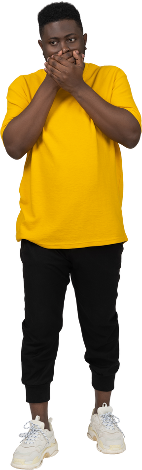 Front view of a shocked young dark-skinned man in yellow t-shirt hiding mouth