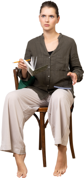 一个困惑的年轻女子穿着家居服坐在椅子上，拿着铅笔和笔记本的前视图