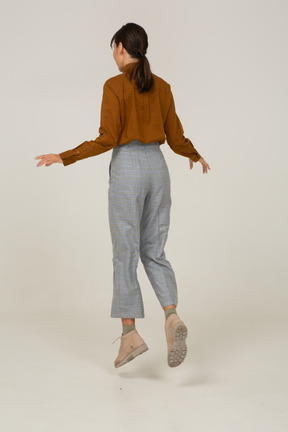 Three-quarter back view of a jumping young asian female in breeches and blouse outspreading hands