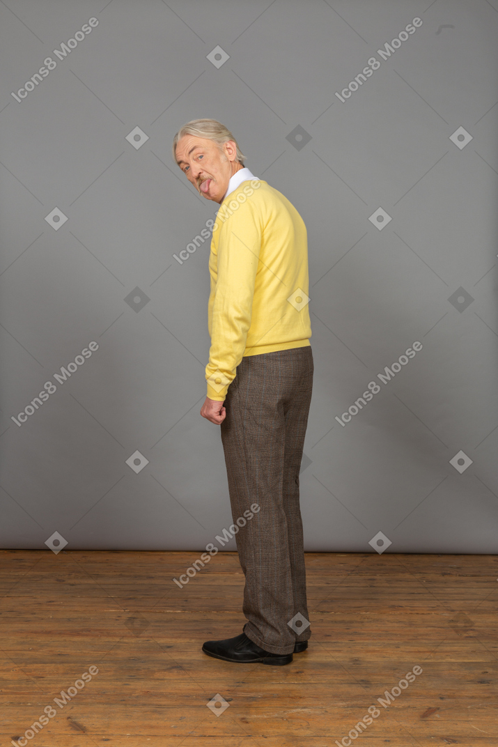 Three-quarter back view of an old curious man in yellow pullover showing tongue and looking at camera