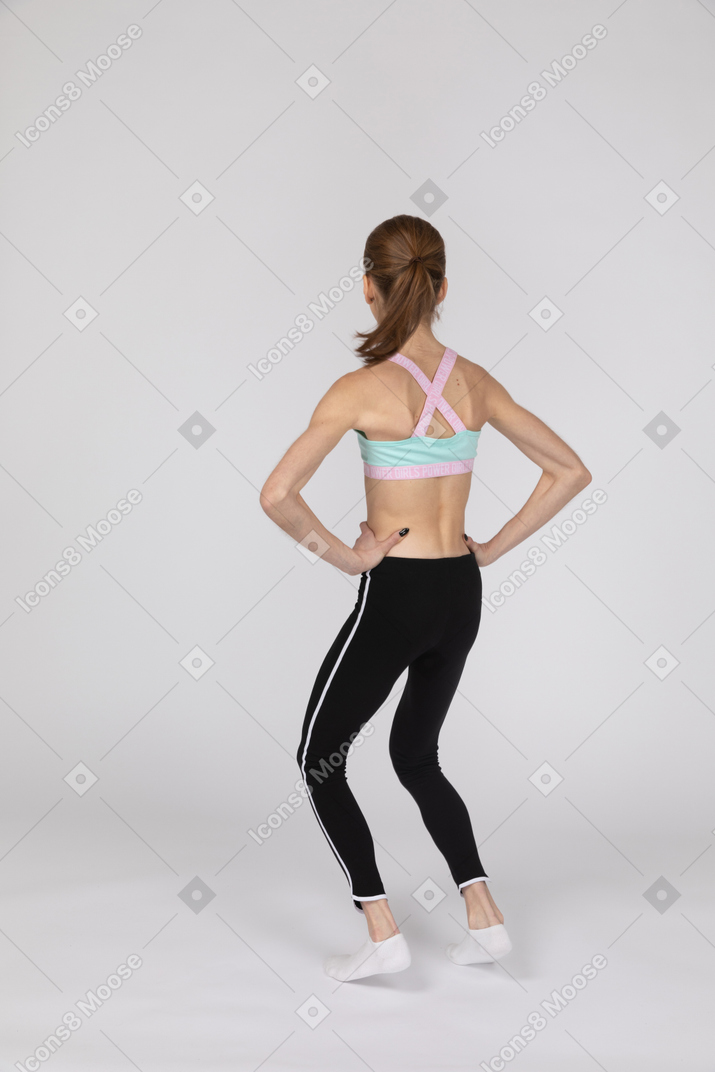 Vista traseira de uma adolescente em roupas esportivas colocando as mãos nos quadris e dobrando os joelhos