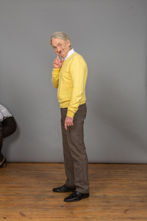 Side view of a cheerful old man in a yellow pullover looking at camera and touching face