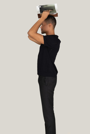 Standing in profile young man in black pants and t-shirt holding magazines on head