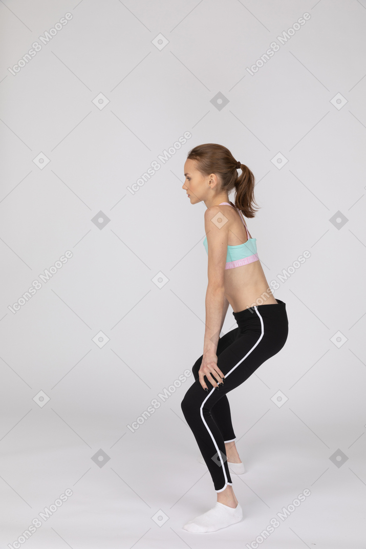 Vista lateral de una jovencita en ropa deportiva en cuclillas y poniendo las manos en las caderas