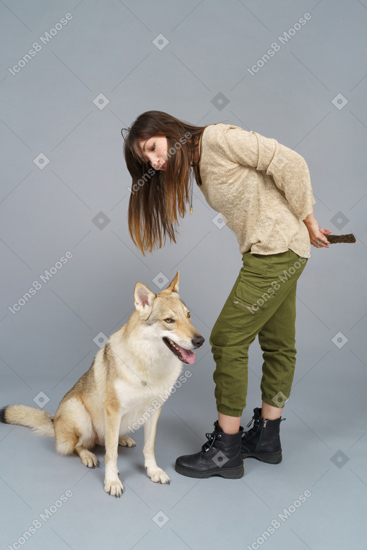 Longitud total de una hembra joven inclinada sobre su perro y sosteniendo un bocadillo detrás de la espalda