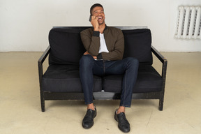 Front view of a smiling young man sitting on a sofa and talking on his phone