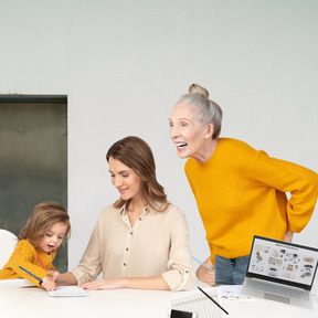 A mom helping a child draw and a grandma standing next to them