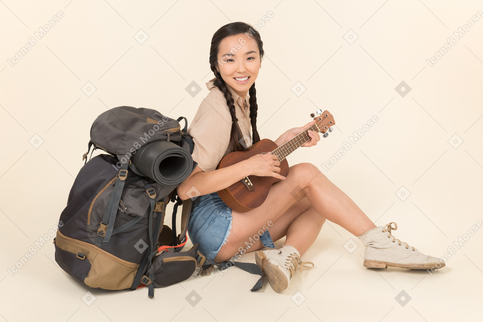 Sonriente joven asiática sentada cerca de la mochila y tocando la guitarra