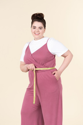 Jovem plus size em um macacão rosa, posando com uma fita métrica contra um fundo amarelo pastel