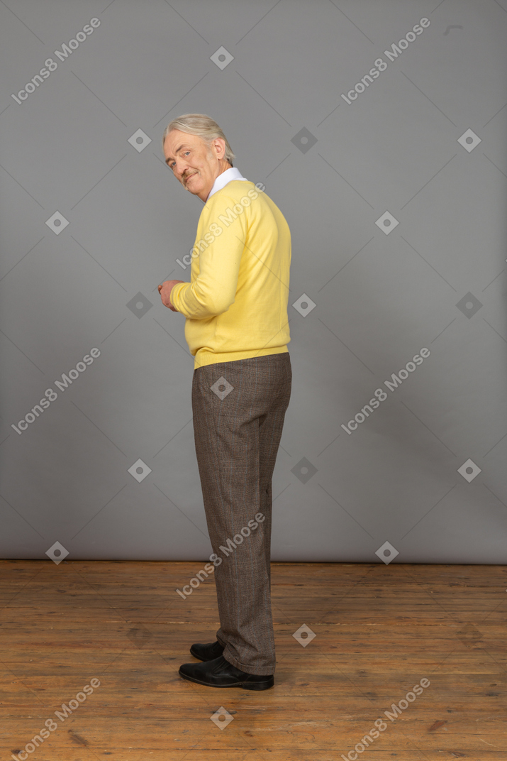 Vista traseira a três quartos de um homem idoso sorridente, vestindo um pulôver amarelo e olhando para a câmera