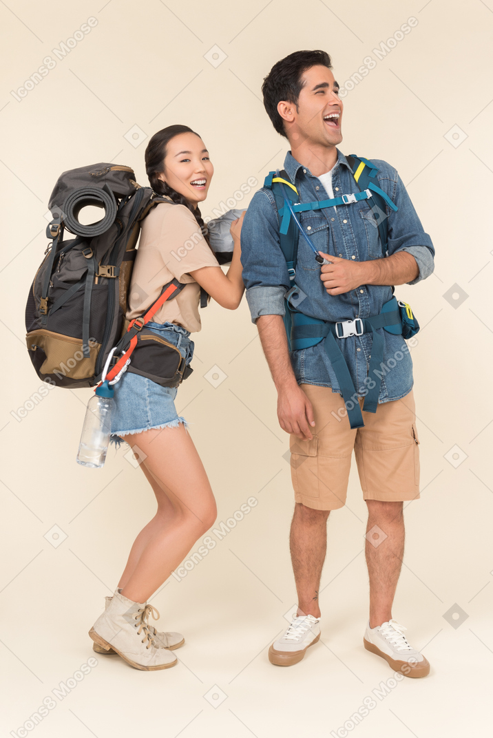 Laughing young asian woman taking something off from his boyfriend's backpack