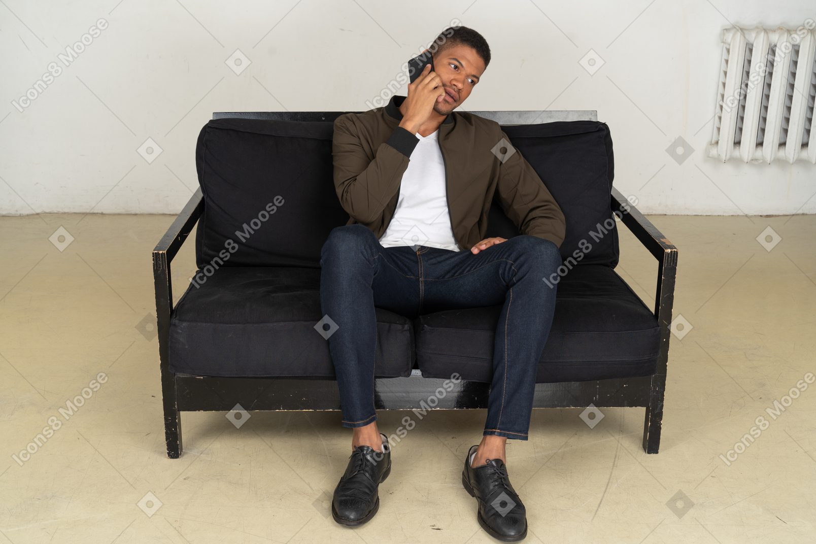 Front view of a perplexed young man sitting on a sofa and talking on his phone