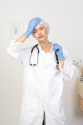 Vista frontal de una joven doctora con estetoscopio sosteniendo el termómetro y tocando la cabeza