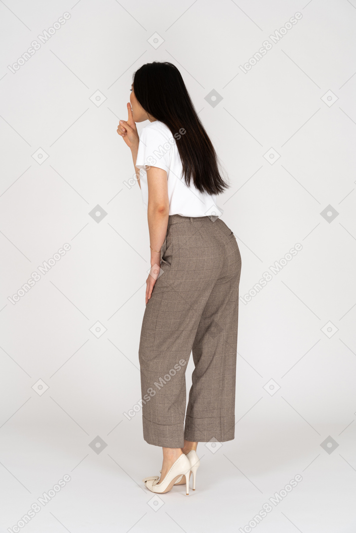 Vista posteriore di tre quarti di una giovane donna in calzoni e maglietta che mostra un gesto di silenzio