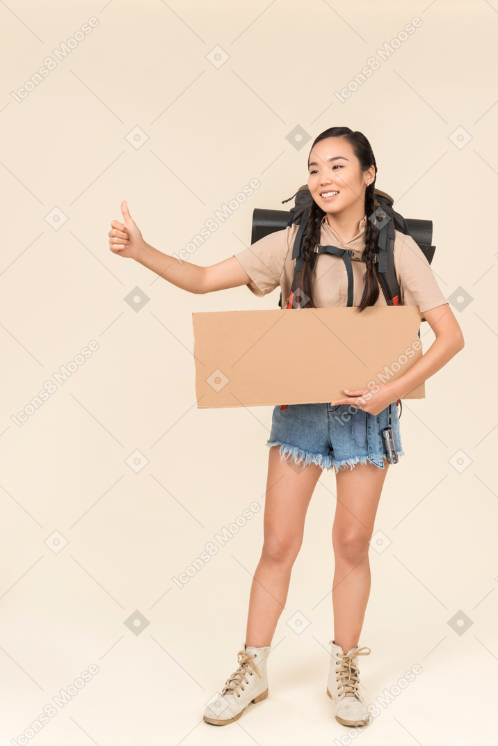 Young female hitchhiker holding paper card and showing thumb up