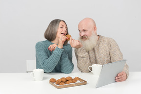 Gealterte paare, die tablette betrachten und kaffee und plätzchen trinken