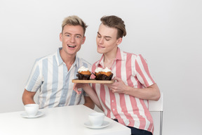 Gay couple sitting at the table and holding tray with cupcakes