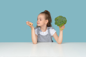 Menina bonitinha tentando escolher entre um donut e alguns brócolis