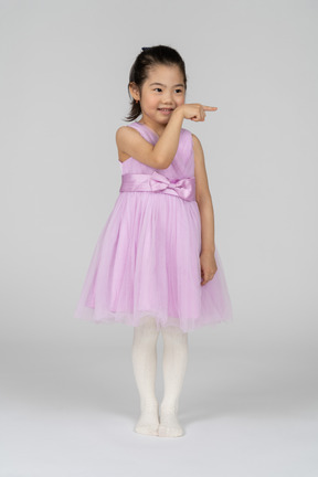 Вид спереди на маленькую девочку в платье-пачке, улыбающуюся, указывая влево