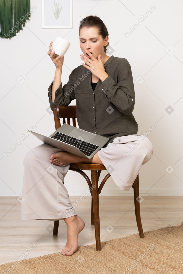 Vista frontal de una mujer joven sorprendida vistiendo ropa de casa sentado en una silla con una computadora portátil y tomando café