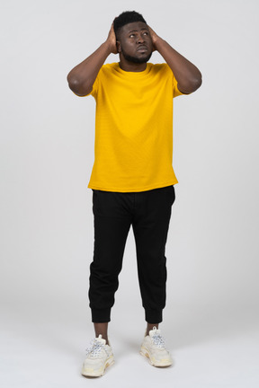 Front view of a young dark-skinned man in yellow t-shirt touching head & looking up
