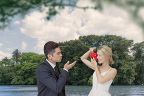 Eine glückliche braut macht ein foto von ihrem bräutigam, der ihr unter den grünen bäumen in der nähe des flusses einen kuss zuwirft