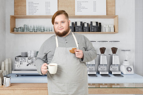 A male barista holding a cup and a cookie in a coffee shop