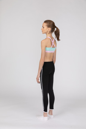 Three-quarter back view of a teen girl in sportswear standing still and looking aside