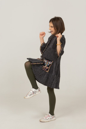 Vista lateral de uma menina feliz em um vestido cerrando os punhos e levantando a perna