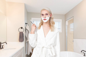 A woman in a bathrobe shaving her face with a razor