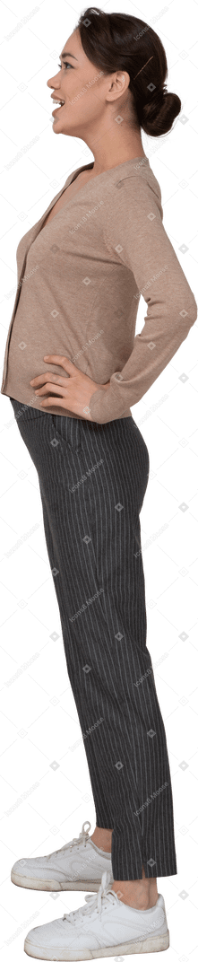 Vue latérale d'une jeune femme souriante en pull et pantalon mettant les mains sur les hanches