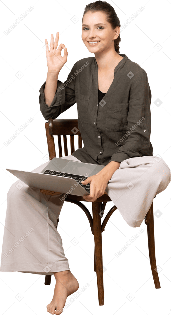一位微笑的年轻女子坐在椅子上，拿着笔记本电脑，摆出 ok 手势的前视图