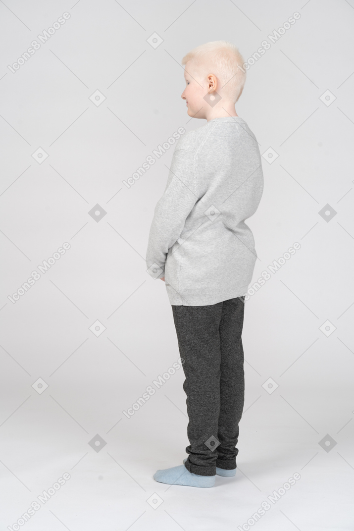 Full-length little blonde boy in casual clothes standing turning back