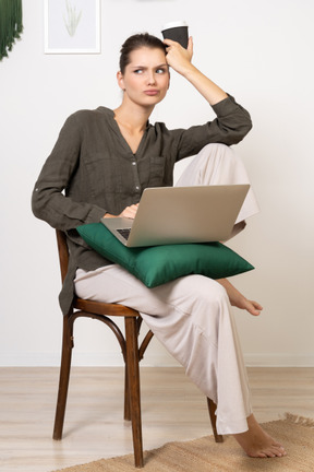 Front view of a confused young woman wearing home clothes sitting on a chair with a laptop and coffee