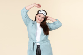 Young plus size woman in a blue bathrobe and a sleep mask on standing against a pastel yellow background, after just waking up