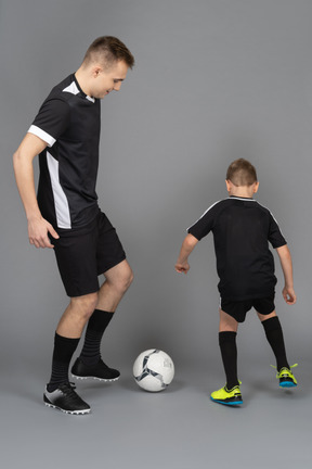 В полный рост молодой человек тренирует маленького мальчика, как играть в футбол