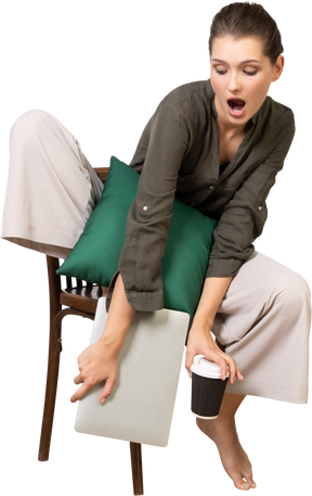 Vista frontal de una mujer joven sorprendida sentada en una silla y sosteniendo su computadora portátil y tocando la taza de café