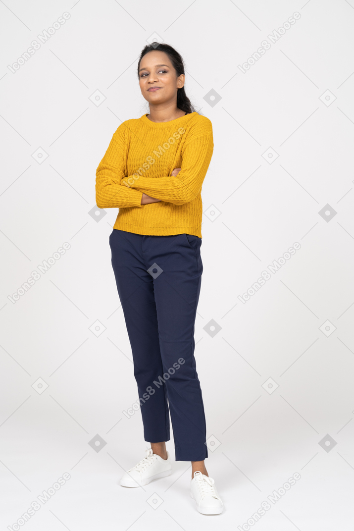 Vista frontal de una niña feliz en ropa casual de pie con los brazos cruzados y mirando hacia arriba