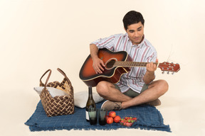 Junger kaukasischer kerl, der nahe picknickkorb auf der decke sitzt und gitarre spielt