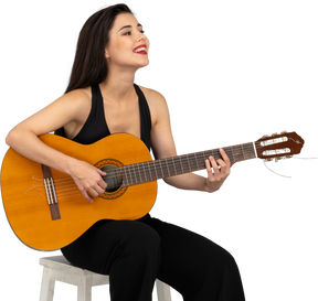Vista de tres cuartos de una joven sonriente sentada en traje negro tocando la guitarra