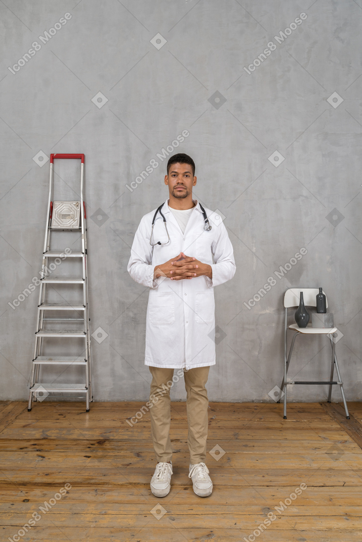 Vista frontal de un joven médico de pie en una habitación con escalera y silla