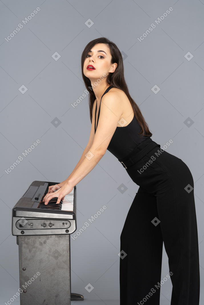 Side view of a young lady in black jumpsuit playing the piano while raising head while looking at camera