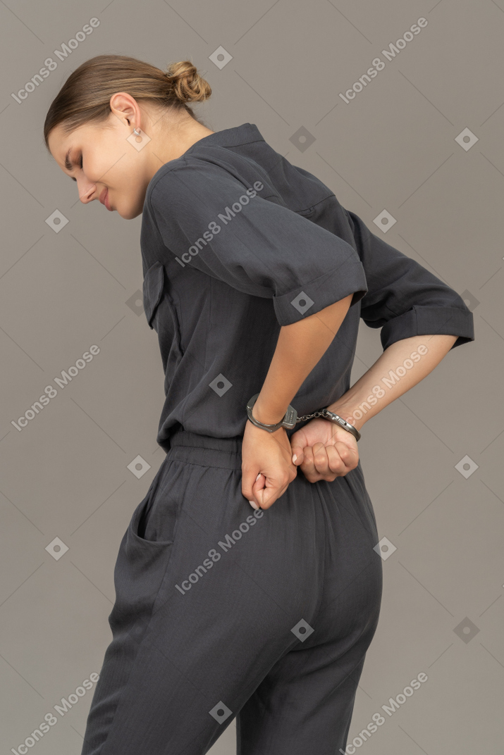 Three-quarter back view of a suffering young woman in a jumpsuit wearing handcuffs