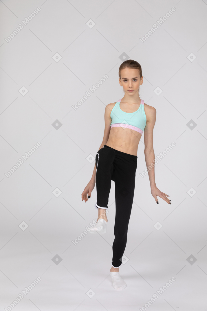 Vista frontal de una jovencita en ropa deportiva extendiendo las manos y levantando la pierna