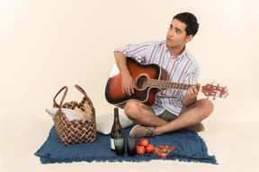 Chico caucásico joven sentado cerca de la cesta de picnic en la manta y tocando la guitarra