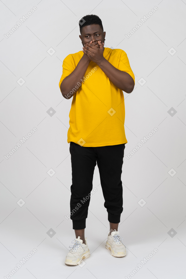 Front view of a shocked young dark-skinned man in yellow t-shirt hiding mouth